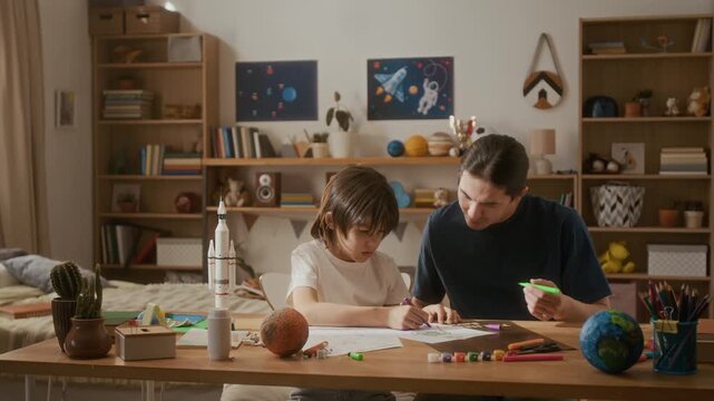 Young adult Asian father helping young multiethnic kid draw space pictures at desk in bedroom. Perfect for family bonding, homeschooling, and creative childhood education commercials