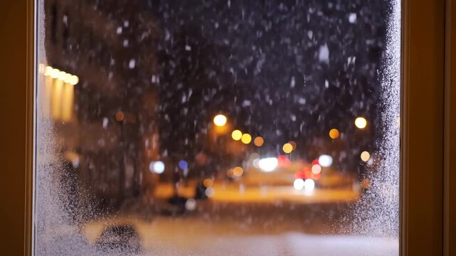 Snow falling outside a window at night with blurred city lights