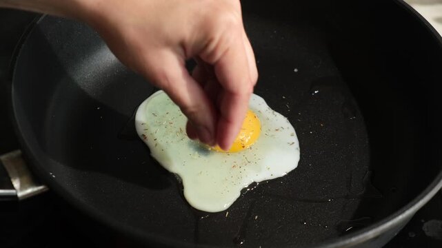 Fried eggs are cooked in frying pan. Female hands sprinkle salt on egg. Preparing morning breakfast, daily routine