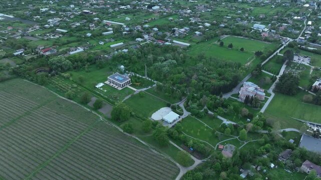 Wide aerial view of Chateau Mukhrani vineyard and estate in Georgia, geometric rows of green grapevines stretching toward the royal palace and village countryside under sunny sky.