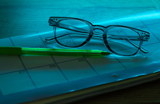 Glasses and a pen on a calendar on a dark desk with subtle blue background lighting. Concept of planning, organization, time management, studying, and working at night