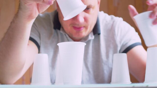 Close-up of hands stacking plastic cups into a fragile tower. Blurred male silhouette. Shallow depth of field, natural light. Metaphor for risk, business growth, stability and delicate balance.