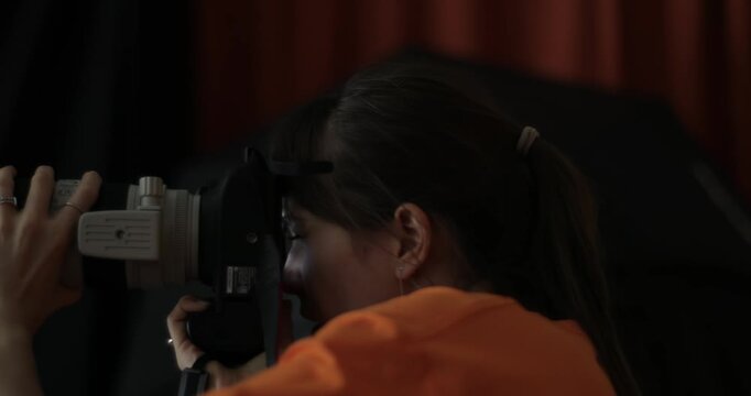Cinematic close-up of young woman in orange top looking through modern camera with telephoto lens in dark studio setting. Perfect for photography, creative, media, and professional content.
