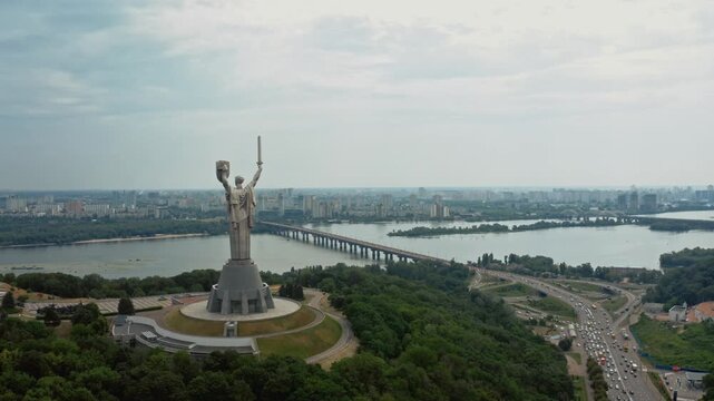 Wide cinematic aerial drone shot of towering Motherland Monument statue dominating Kyiv skyline above the Dnipro River with bridge and city in background. Perfect for Ukraine, travel, and landmark con
