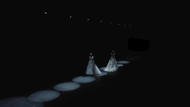 Wide aerial shot of two elegant models in white wedding gowns walking along dark runway illuminated by circular LED ring lights at bridal fashion show. Perfect for wedding, fashion, and luxury content