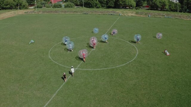 Wide aerial drone shot of group playing bubble soccer in colorful inflatable zorb balls on green grass field surrounded by trees. Perfect for fun, sport, entertainment, and team event content