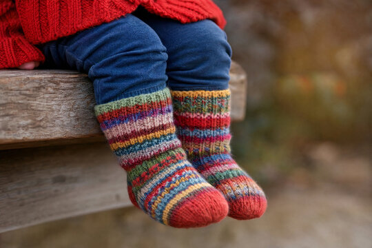 Close up of a toddler sitting on a rustic wooden bench wearing bright multicolored striped knitted wool socks and a red sweater outdoors during autumn.