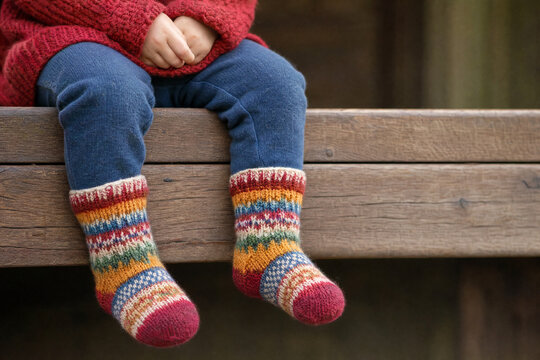 A toddler sitting on a weathered wooden bench wearing colorful hand-knitted wool socks with traditional patterns and a red sweater for a cozy autumn lifestyle concept.