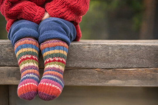 A young child wearing colorful striped wool socks and a red knit sweater sits on a rustic wooden bench outdoors, representing cozy winter fashion and childhood warmth.