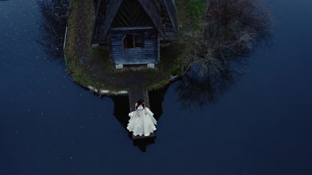 Close aerial drone shot looking down at elegant bride in white wedding dress standing beside old abandoned wooden boathouse on small island in calm blue lake. Perfect for wedding and cinematic content