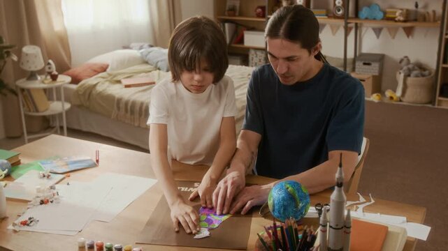Asian father and young multiethnic son gluing paper astronaut onto poster at desk, discussing space with globe and rocket model nearby. Footage ideal for homeschooling and STEM education commercials