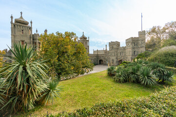 Medieval-style Massandra Palace with multiple towers and turrets set against backdrop of greenery. Palm-like plants frame foreground while autumn-colored trees add warmth to scene. Yalta, Crimea © SergioDenisenko