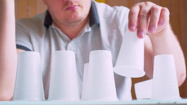 Man's hands building a pyramid with plastic cups. Blurred silhouette in a  room. Close-up, shallow depth of field, natural warm light, cinematic footage.