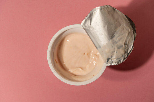 Open plastic yogurt cup on pink background top view. Top view of an open round plastic tub of fresh peach fruit yogurt with peeled back foil lid on a pink surface