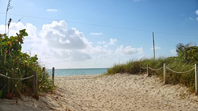 Sandy path to the ocean through coastal dunes, Miami Beach, Florida. Rope fence lines lead toward turquoise water under partly cloudy sky.