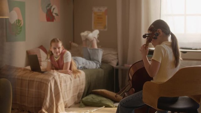 Handheld shot of Caucasian child playing cello while teenager lies on bed looking at laptop during remote music lesson. Ideal for e-learning and home education marketing campaigns