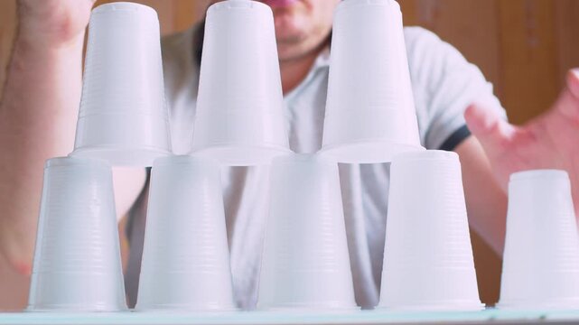 Man's hands dismantling a plastic cup pyramid. Blurred silhouette in a room.Close-up, shallow DoF, natural light, cinematic bokeh.Metaphor for safe deconstruction, project completion and risk control