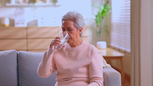 Senior woman drinking water from glass on sofa in bright living room, hydration and wellness routine for elderly female at home interior, calm morning healthy lifestyle and self care