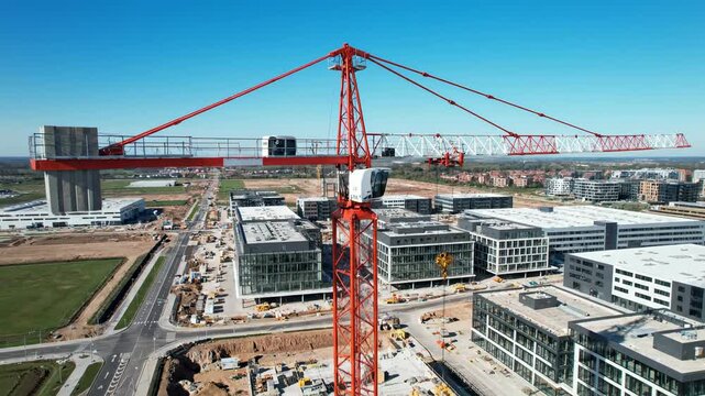 Majestic red tower crane dominates a sprawling construction site with several impressive modern buildings under development beneath a vast, clear blue sky