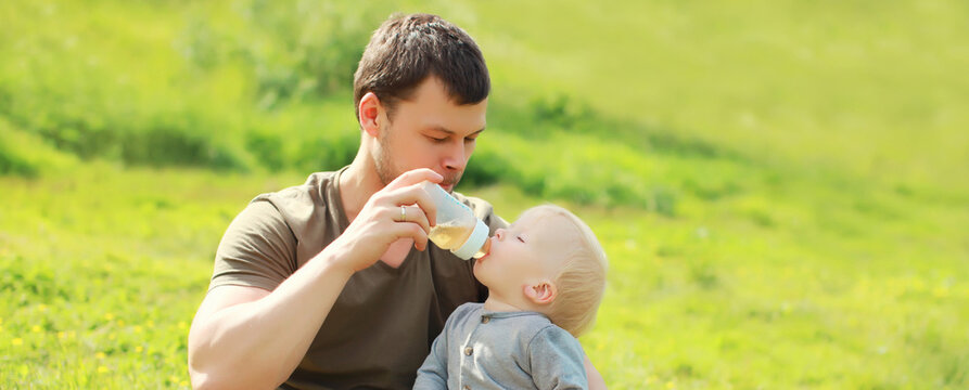 Father feeds baby from bottle sitting on grass in sunny summer park