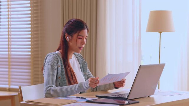 Working woman in home office using laptop for online meeting, earbud on, reviewing document during remote video call at desk, calm professional remote work setup, focused woman at home office desk
