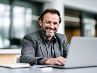 Fototapeta premium Smiling mature man in a gray blazer working on a laptop in a bright, modern office with a notebook and smartphone nearby.
