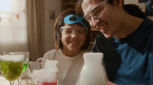 Multiethnic child and Asian man wearing safety goggles watching dry ice chemical reaction with colorful liquids. Ideal for STEM education and family learning advertisements