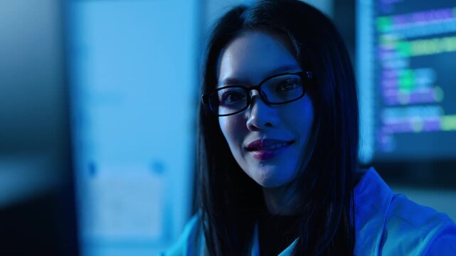 Smiling Asian young female IT professional or programmer wearing glasses and working in a dark server room with blue lighting. Computer monitors displaying code in background. Cybersecurity concept.