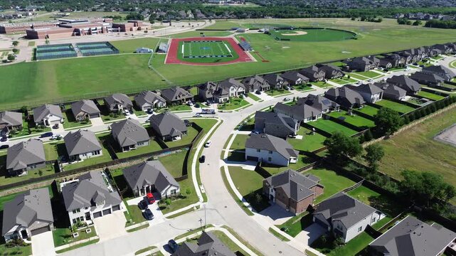 Campus facing neighborhood in Arlington, TX displays rows of homes with fenced yards, parked cars clean streets across from athletic fields academic buildings, upscale master planned community