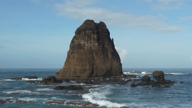 Cinematic shot of giant sea stack rock formation in the middle of blue ocean with waves crashing against the rocky coast under bright sunny sky. Papuma Beach, Jember, Indonesia
