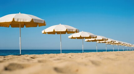 Fototapeta premium A row of white and yellow beach umbrellas on a sandy shore under a clear blue sky.