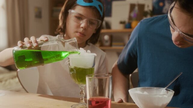 Tilt up view of Asian man and multiethnic child wearing safety goggles conducting chemistry experiment, pouring green liquid into glass with dry ice. Clip ideal for promoting science kits