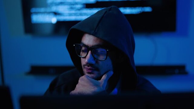 Focused young male hacker in a dark hoodie and glasses works intently on a laptop in moody blue-lit room. Complex coding displays on the background screen. Perfect for cybersecurity and tech concepts