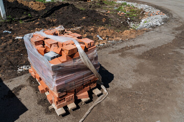 Fototapeta premium Pallet Of Bricks Roadside Assembly. Urban DIY Project With Chipped Brick Units. Masonry Supplies Consisting Of Red Bricks On Pallet With Damaged Wrapping Ready For Building Repairs