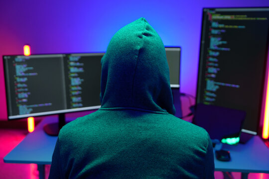 Hooded hacker coding at dual monitors in neon lit room