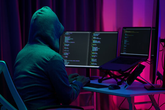 Hooded Hacker Coding at Dual Monitors in Dark Neon Room