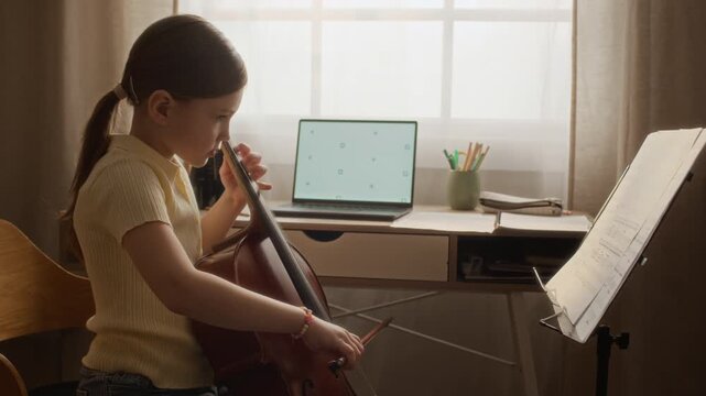 Side view of Caucasian child practicing cello during online music lesson with laptop showing mockup screen. Clip suitable for promoting remote education, e-learning platforms, and music tutoring apps