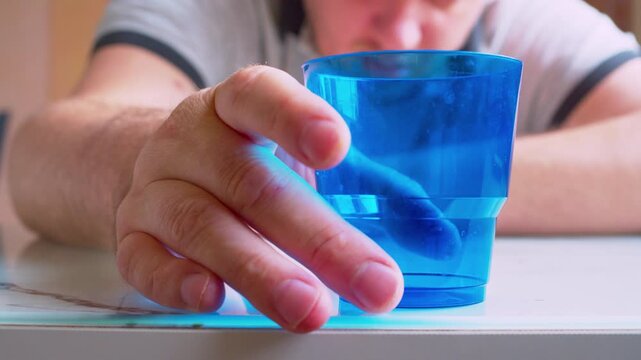 Close-up of a hand placing an empty plastic cup on a table, blurred silhouette of a drunk man swaying and falling asleep background.Shallow depth of field.Cinematic shot of an intoxicated swaying man