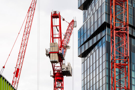 Red construction crane rises between glass building and office facade, showing machinery, vertical frame and urban development at close range