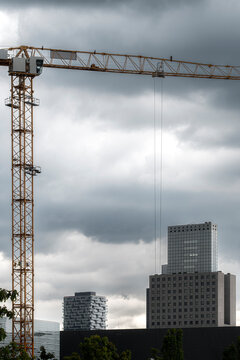 Tower crane stands against storm clouds above downtown buildings, with suspended hook, skyline layers and construction site tension in the distance
