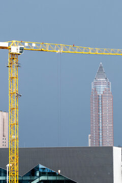Yellow construction crane spans across a city skyline beside a skyscraper, tower, roofline and glass facade under clear blue sky above