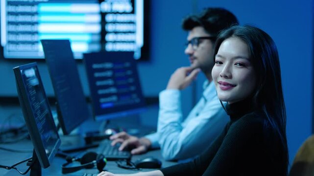Smiling Asian young female IT developer looking at camera while working on computer with male colleague in modern blue-lit control room, cyber security software engineer team operating data center