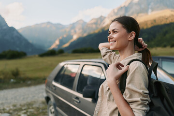 Fototapeta premium A smiling woman in casual clothes stands near a car in a mountain landscape, enjoying the outdoors and fresh air. Concept for lifestyle, travel, and freedom.