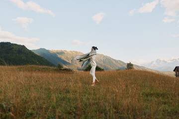 Fototapeta premium Young woman in casual clothes enjoys freedom and fresh air running through grassy field with mountains in background under blue sky with clouds.