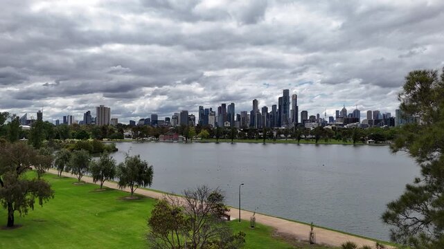 Smooth cinematic drone pan across Albert Park Lake, revealing the Melbourne skyline and modern urban architecture under dramatic cloudy skies.