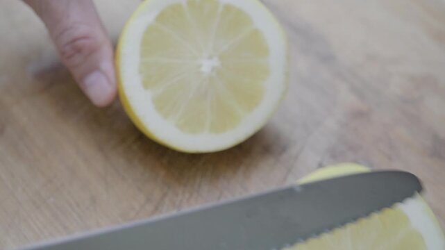 Hand slicing fresh ripe lemon with sharp knife on wooden cutting board. Juicy citrus rounds reveal pulp detail in natural light. Cooking and healthy food preparation concept.
