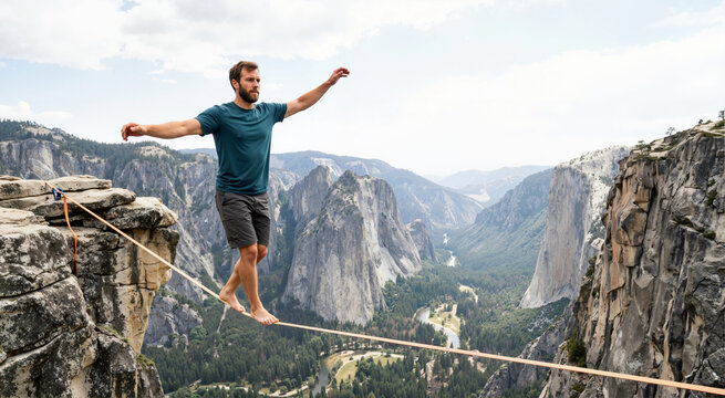 Brave man balancing on slackline high above scenic valley. Male athlete performing extreme sport on highline in mountains