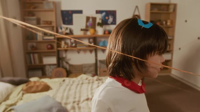 Multiethnic child and Asian father wearing superhero capes playing laser maze game with yarn in bedroom. Ideal for family lifestyle, parenting, and creative play promotions