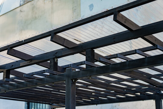 Modern outdoor wooden pergola structure with translucent corrugated plastic roofing sheets installed on dark painted timber beams for backyard patio construction