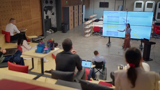 A female professional presents project timelines to colleagues in an amphitheater-style meeting space. Diverse employees attentively listen to a presentation on a large screen